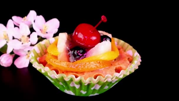 Cream cupcake with cherry, berry, fruits and apple flowers on black background rotate slowly. — Stockvideo