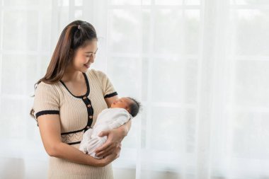 Güzel bir Asyalı kadın kucağında bebek kucağıyla evinin penceresinin önünde duruyor. Mutlu bebek annesinin kollarında ve rahat bir şekilde uyuyor. Anne ve bebek kavramı.