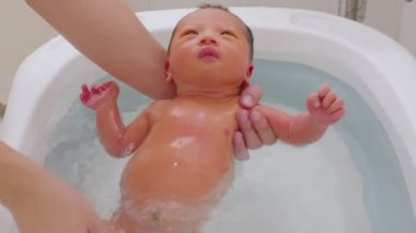 Asyalı yenidoğan bebeğin küvette banyo yapması. Anne oğlunu ılık suda yıkıyor. 7 gün boyunca yeni doğan bebek gülüşü küvette rahatlıyor ve rahatlıyor.