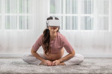 Mutlu Asyalı hamile kadın sanal gerçeklik gözlüklerini pratik için kullanıyor yoga sınıfı kelebeği, meditasyona büyük göbekli rahatla evde. Hamilelik Sanal Gerçeklik Teknolojisi Konsepti