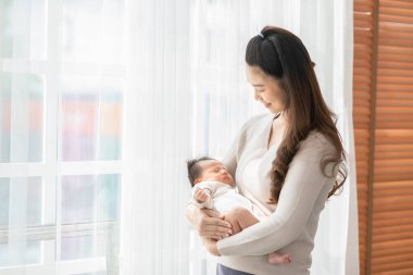 Güzel bir Asyalı kadın kucağında bebek kucağıyla evinin penceresinin önünde duruyor. Mutlu bebek annesinin kollarında ve rahat bir şekilde uyuyor. Anne ve bebek kavramı.