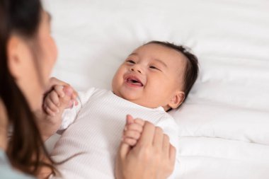 Yeni Asyalı anne yatakta sevimli yeni doğmuş bebeğe oynuyor gülümseyerek ve evde mutlu bir şekilde. Anne bebek bebekle konuşuyor ve el ele gülüyor. Bebek ve Anneler Günü konsepti.