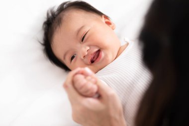 Yeni Asyalı anne yatakta sevimli yeni doğmuş bebeğe oynuyor gülümseyerek ve evde mutlu bir şekilde. Anne bebek bebekle konuşuyor ve el ele gülüyor. Bebek ve Anneler Günü konsepti.
