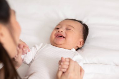 Yeni Asyalı anne yatakta sevimli yeni doğmuş bebeğe oynuyor gülümseyerek ve evde mutlu bir şekilde. Anne bebek bebekle konuşuyor ve el ele gülüyor. Bebek ve Anneler Günü konsepti.