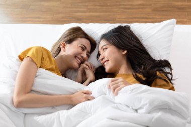 Güzel Asyalı genç lezbiyen çift ya da yatağa uzanmış birlikte gülümseyen ve sevgiyle rahatlayan arkadaşlar. Mutluluk LGBTQ çifti evde birlikte vakit geçiriyor. LGBTQ Yaşam Biçimi Kavramı