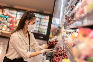 Tıbbi maske takan güzel çekici Asyalı kadın ve lastik eldivenler süpermarketteki meyve raflarından üzüm seçiyor. Genç bir bayan bakınıyor ve markette üzüm alışverişi yapıyor.