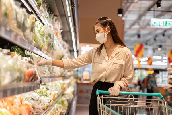 Tıbbi maske takan güzel bir Asyalı kadın ve lastik eldiven süpermarkette sebze ve meyve seçen bir kadın. Markette kadın arıyor ve sebze alışverişi yapıyor.