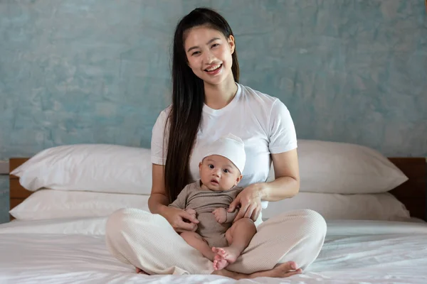 白い居心地の良い家でベッドに座っている彼女の腕の中で新生児を保持する美しいアジアの母の女性 お母さんの腕の安全性と快適で幸せな赤ちゃんの笑顔 お母さんと赤ちゃんのコンセプト ロイヤリティフリーのストック画像