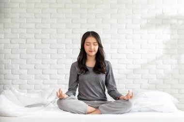 Sağlıklı Asya 'lı genç bir kadın yatakta yoga nilüferiyle soluk alıp veriyor. Yoga yapıyor, sağlıklı genç bir kadının meditasyonunu yapıyor. Beyaz rahat evinde dinleniyor ve rahat ediyor.