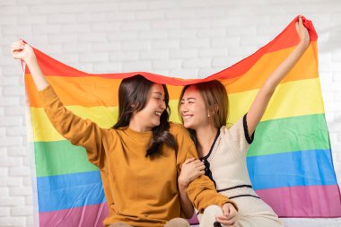 LGBTQ Asyalı genç lezbiyen çift, birlikte gururla bayrak mutluluğuna gülümsüyor. Neşeli homoseksüel çift, LGBTQ gurur bayrağı altında kucaklaşıyor ve kameraya bakıyor. LGBTQ Yaşam Tarzı Onur Ayı