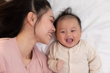 Yeni Asyalı anne yatakta sevimli yeni doğmuş bebeğe oynuyor gülümseyerek ve evde mutlulukla. Anne bebekle konuşuyor ve bebek yanağından sevgiyle öpüyor. Bebek ve Anneler Günü konsepti.