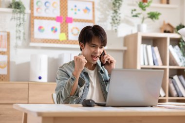 Girişimci yakışıklı Asyalı adam laptop ile çalışırken günlük mavi tişört giyer ve ev ofisinde kutlama yapan cep telefonuyla müşteri veya iş ekibiyle konuşur. İş Kuruluşu Konsepti