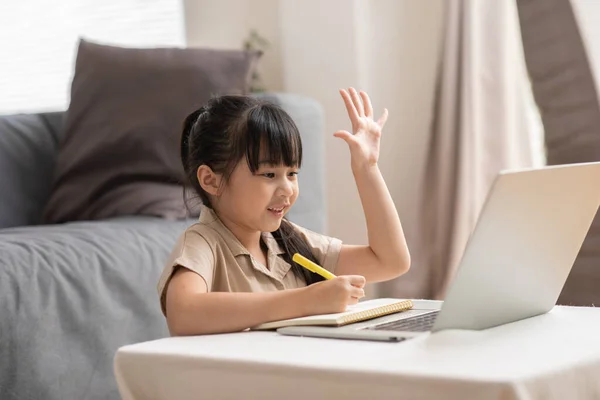 小さなアジアの女の子の子供は 家庭の笑顔と幸福でオンラインクラスでの質問に答えるために手を上げるコンピュータのラップトップ上でオンライン学習を勉強します ロイヤリティフリーのストック画像