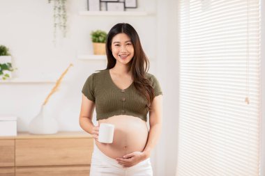 Pencerenin önünde duran mutlu hamile kadın beyaz fincanı tutuyor ve büyük karnını sevgiyle okşuyor. Genç bir kadının hamileliği gelecekteki hayattan zevk alıyor. Aşkla rahatla. Annelik ve hamilelik.
