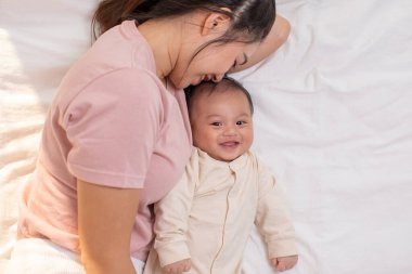 Anne ve çocuk uzanıyorlar ve sarılıyorlar. Anne oğlunu öpüyor ve ikisi de gülümsüyor. Sevimli erkek bebek çok mutlu görünüyor ve anneyle oynamaktan zevk alıyor. Bebek tombul ve sağlıklı..