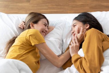 Güzel Asyalı genç lezbiyen çift ya da yanaklarına dokunan ve birlikte gülümseyip yatağa uzanan arkadaşlar. Mutluluk LGBTQ çifti evde birlikte vakit geçiriyor. LGBTQ Yaşam Biçimi