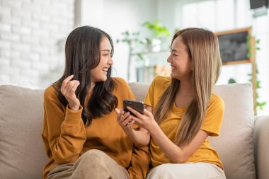 Genç Asyalı lezbiyen çift oturma odasındaki kanepede dinlenirken dijital cihaz kullanarak eğleniyor. İki eşcinsel kadın ekranda sörf yapmaktan zevk alıyor. Video izliyorlar.. 