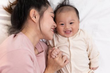 Anne ve çocuk uzanıyorlar ve sarılıyorlar. Anne oğlunu öpüyor ve ikisi de gülümsüyor. Sevimli erkek bebek çok mutlu görünüyor ve anneyle oynamaktan zevk alıyor. Bebek tombul ve sağlıklı..