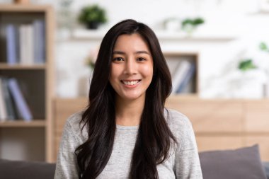 Çekici, Çekici, Asyalı genç bir kadın kanepeye oturup kameraya bakar ve evde mutlu ve rahat bir şekilde gülümser. Sevimli kız kolları çapraz güvenle güler ve evdeki oturma odasında rahatlar.