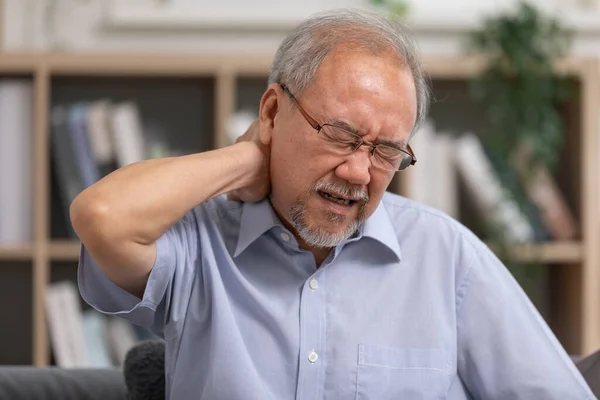 Asian Older man suffering from shoulders and neck pain sitting on sofa. Health problem retired Asian old man hands touching and massage neck for relief pain . Healthcare concept, elderly care.