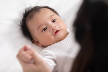 Yeni Asyalı anne yatakta sevimli yeni doğmuş bebeğe oynuyor gülümseyerek ve evde mutlu bir şekilde. Anne bebek bebekle konuşuyor ve el ele gülüyor. Bebek ve Anneler Günü konsepti.