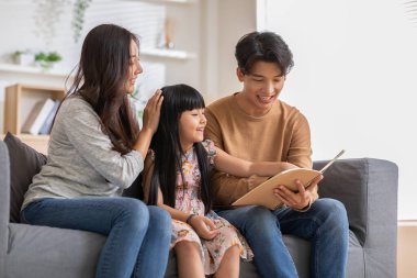Mutlu Asyalı aile kanepede oturup kızının not defterine bakıyor. Ebeveynler gülümsüyor, kızlarının kitabını okuyor ve eğitimle gurur duyuyorlar. Birlikte kitap izlemek, aile ilişkisi.