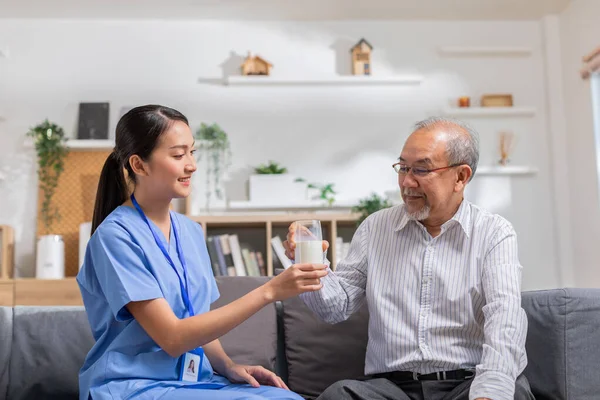Asyalı güzel terapist doktor huzurevindeki yaşlı hastaya süt veriyor. Mavi üniformalı Asyalı hemşire yaşlı bir adama koltukta otururken süt içmesi için yardım ediyor.