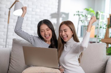 Mutlu genç Asyalı çift dizüstü bilgisayar kullanarak kanepede oturup video izliyor. Her ikisi de ekrana bakıp gülerek cihaza iyi haberi veriyor. LGBT konsepti ve LGBT topluluğu.