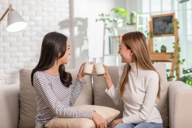 Genç Asyalı kadın çift birlikte organik taze süt içiyor. LGBTQ çifti ellerinde süt tebessümü ve evde kanepede sağlam bir kemik için birlikte kıkırdıyor.