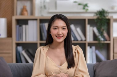 Çekici, Çekici, Asyalı genç bir kadın kanepeye oturup kameraya bakar ve evde mutlu ve rahat bir şekilde gülümser. Sevimli kız kolları çapraz güvenle güler ve evdeki oturma odasında rahatlar.