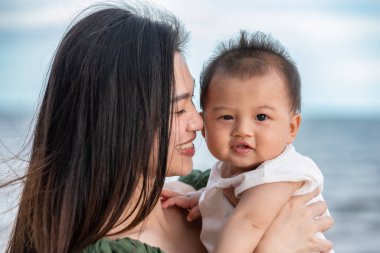 Güzel, Çekici Asyalı anne yeni doğmuş bebeğini elinde tutuyor ve kumsalda bebek başını öpüyor. Mutlu anne ve bebek birlikte gülümsüyor ve gün batımını seyrediyor.
