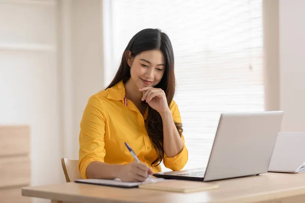Entrepreneur beautiful business asian young woman wear yellow shirt wrinting on paper working online with laptop.Freelance woman working online sale marketing at home.Small Business Startup concept