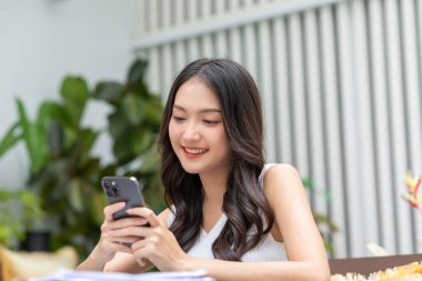 Mutlu Asyalı genç kadın akıllı telefon kullanarak gülümsüyor ve sosyal medyayla veya evde online alışveriş yaparak eğleniyor. Cep telefonu eğlence ve mutluluğuna bakan güzel bir kadının modern sağlık hayatı.