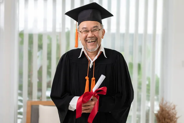 Asian elderly in cap and gown smile and celebration proud success graduate. Cheerful senior man holding certificated or diploma glad and happiness. Elderly graduation concept