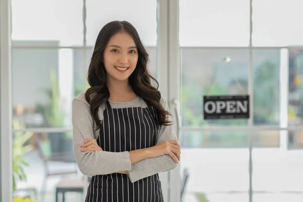 Happy Asian young business owner woman standing amile and cross arm in coffee shop cafe with open label in background. Cheerful of confident entrepreneur female looking at camera. Small business