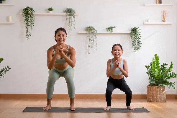 Healthy couple mom and little girl doing yoga squat up and down on mat at home.Mother and female daughter practice yoga breathing exercise and meditate together calm and comfort.Happy Family Activity