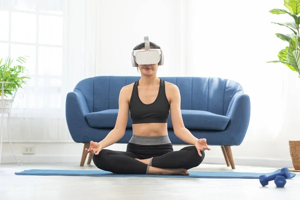 Asian woman using VR virtual reality glasses headset greeting friends in yoga class.Young female goggle using VR enjoy exciting exercise yoga watching 360 degree at home.Virtual Reality Metaverse