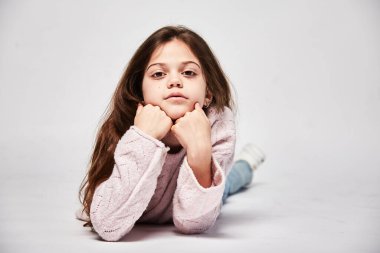 Doğulu görünümlü şirin 10 yaşında bir manken kameraya bakar, kazak ve kot pantolon giyer, reklam kıyafetleri giyer ve gülümsemez. Beyaz bir fotokopi stüdyosunun portresi.