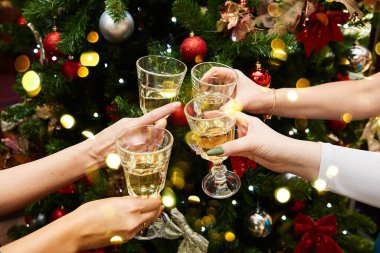 Girls celebrate New Year and Christmas, hands with glasses on the background of the Christmas tree and bokeh. Close-up of a group of people clinking glasses of wine or champagne against a background of bokeh.