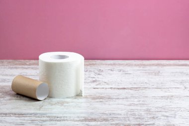Bir rulo beyaz delikli tuvalet kağıdı boş bir rulonun yanında duruyor. Pembe arka plandaki ahşap bir masada.