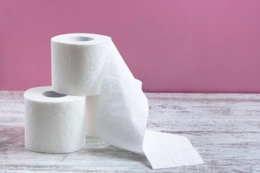 Bir yığın beyaz delikli tuvalet kağıdı, bir rulo açılmamış. Pembe arka plandaki ahşap bir masada.