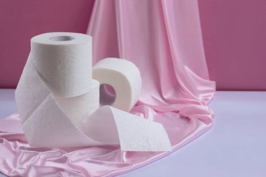 Üç rulo beyaz delikli tuvalet kağıdı, bir tanesi açılmamış, pembe ipek arka planda.