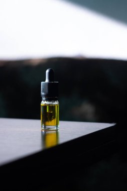 A glass bottle with a pipette filled with cbd oil stands on a dark wood table, against a dark background.
