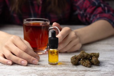 Kadın eli bir fincan çay tutar. Ön planda kenevir cbd yağlı pipetli cam bir şişe ve yakınlarda kuru marihuana tomurcukları var.