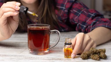 Kadın elleri çaya pipetten marihuana cbd yağı ekler. Yakınlarda çeşitli tıbbi esrar tomurcukları var.