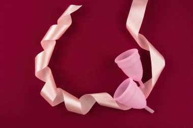 Two eco-friendly, reusable pink colored menstrual cups made of soft medical grade silicone lie flanked by a silk ribbon.  On a dark red blood-colored background