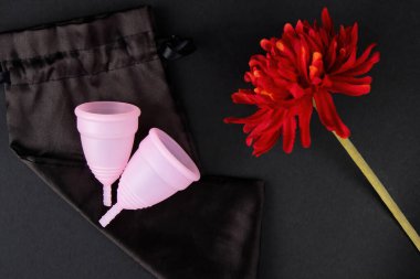İki çevre dostu pembe adet kupası, siyah bir arka planda, kırmızı bir çiçeğin yanındaki ipek bir depoda yatıyor.