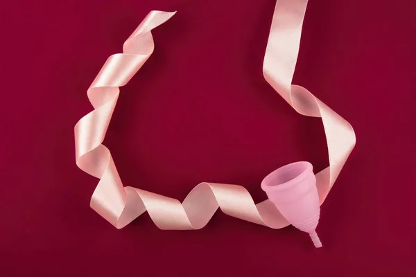 Eco-friendly, reusable pink menstrual cup made of soft medical silicone lies surrounded by a silk ribbon.  On a dark red blood-colored background