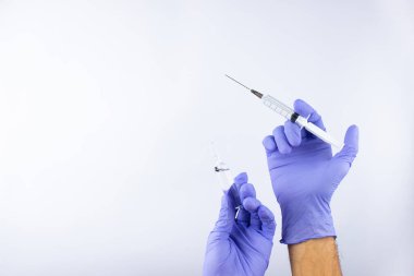 a disposable syringe with a transparent needle and an ampoule of liquid in male hands in blue (lilac) nitrile gloves.  On a white background, lots of empty space
