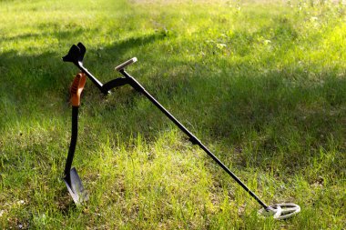 The wireless metal detector is mounted on a shortened metal shovel. Grass and trees in the background